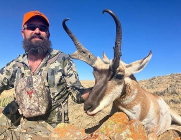 A hunter with a beard in camouflage and an orange cap proudly posing with his trophy antelope in a Wyoming prairie, the clear blue sky enhancing the vibrant colors.